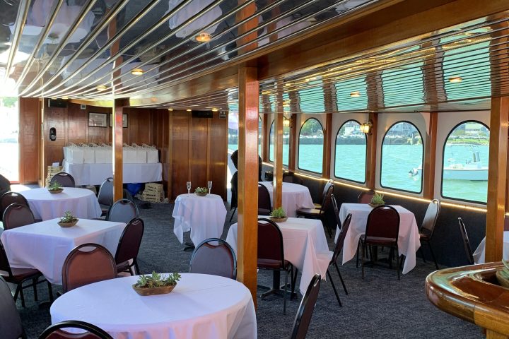 Interior of a boat with round tables, white tablecloths, and chairs, featuring arched windows with water views.