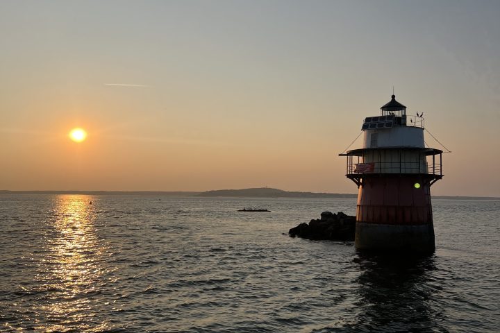 Sunset over ocean with a lighthouse on the right and distant hills on the horizon.