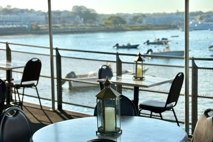 Outdoor cafe with table, chairs, and lantern overlooking a calm bay with boats in the background.