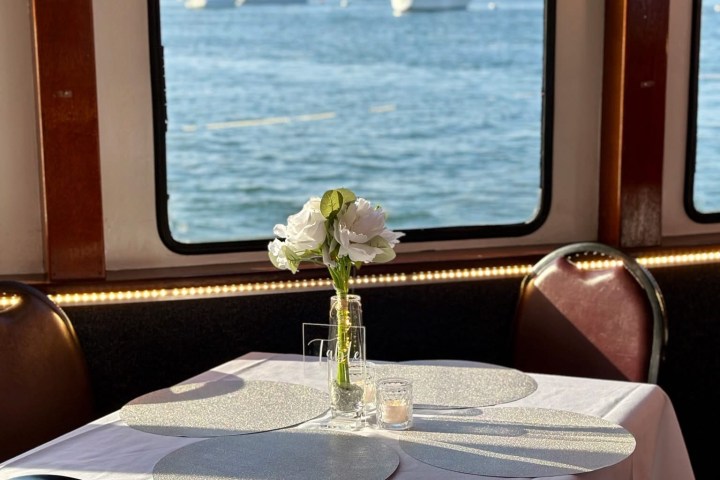 Table with white flowers by a window overlooking a harbor with boats.