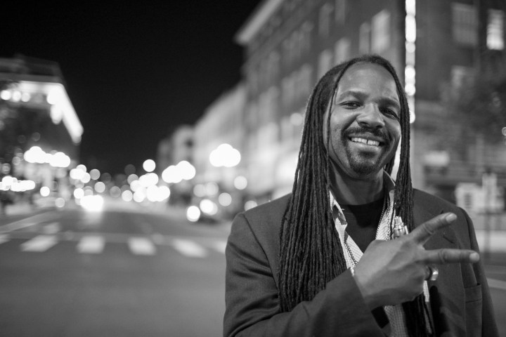 Man with long hair smiles and gestures peace sign on a city street at night.