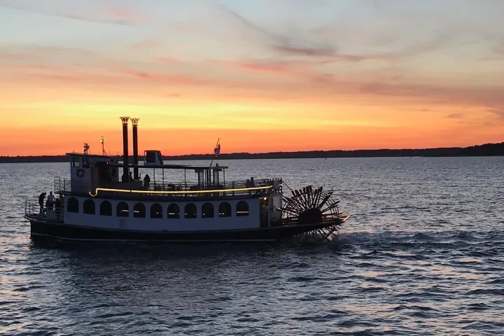 Riverboat cruising on water at sunset with vibrant sky.